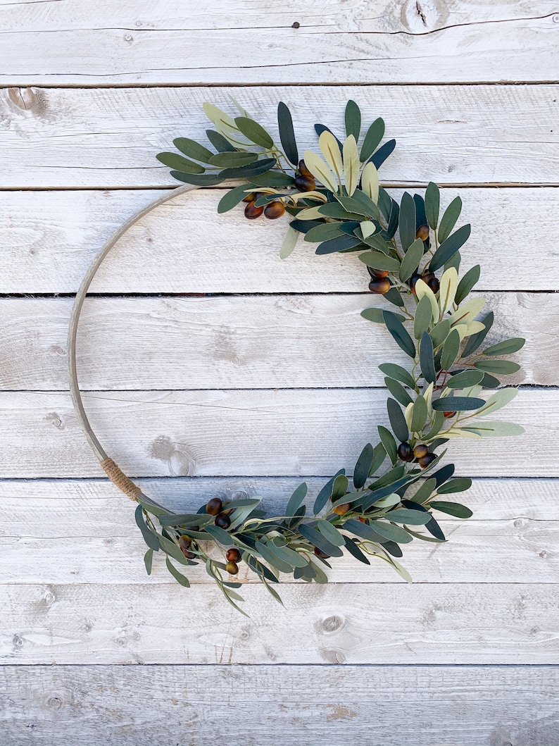 May include: A green olive branch wreath with brown olives, arranged in a circle on a white wooden background.