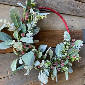 May include: A red wreath with frosted eucalyptus leaves and red berries. The wreath is hanging on a wooden wall.