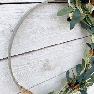 May include: A gray metal wreath form with a brown twine bow. A green olive branch with brown olives is wrapped around the wreath form.