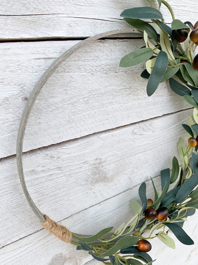 May include: A gray metal wreath form with a brown twine bow. A green olive branch with brown olives is wrapped around the wreath form.