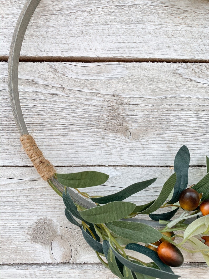 May include: A rustic wooden wreath with a brown twine accent and a green olive branch with brown olives.