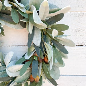 May include: A green and gray olive branch wreath with brown olives. The wreath is made of artificial leaves and is perfect for decorating a home or office.