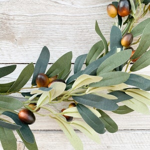 May include: A green olive branch wreath with brown olives. The wreath is on a white wooden background.