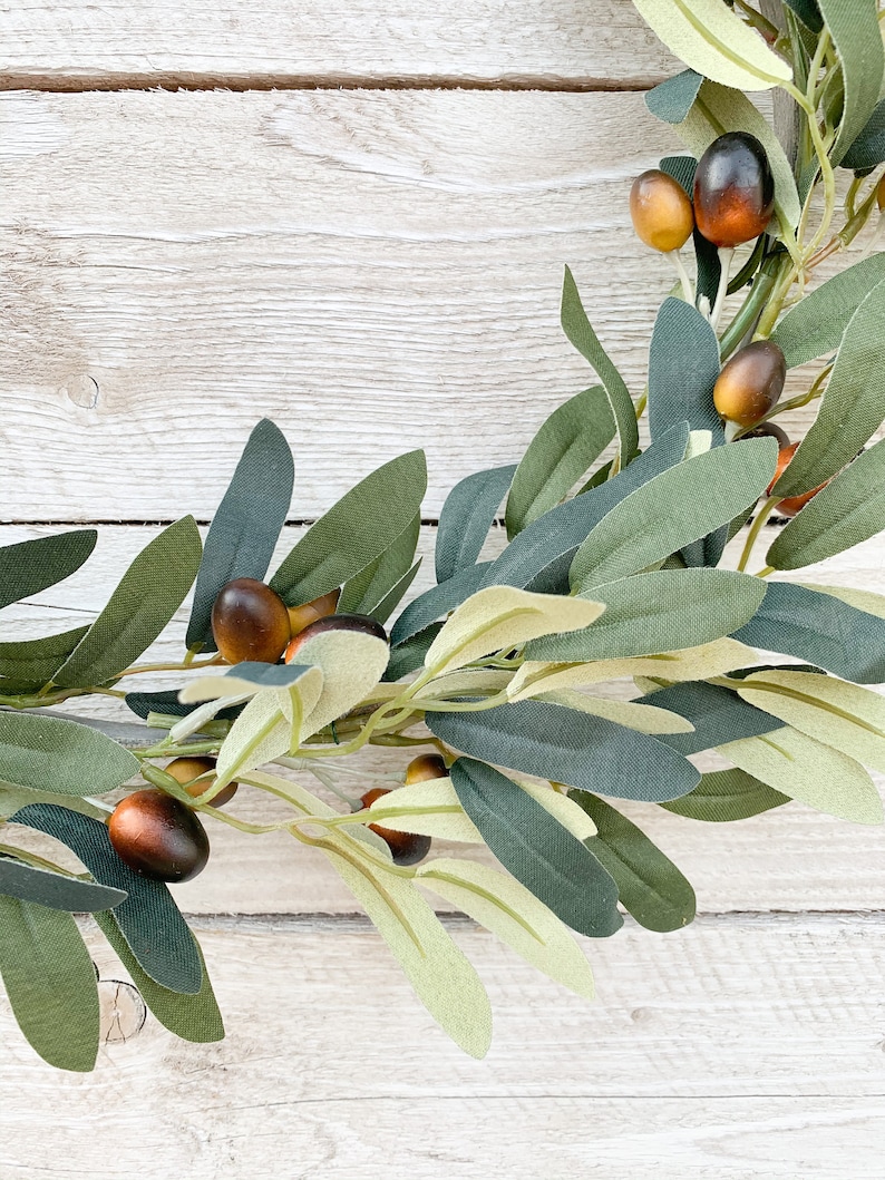May include: A green olive branch wreath with brown olives. The wreath is on a white wooden background.