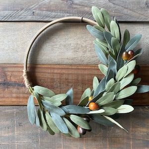 May include: A green olive branch wreath with brown berries, displayed on a wooden background.