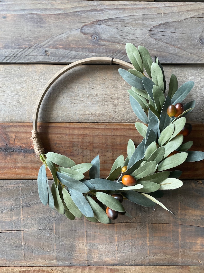 May include: A green olive branch wreath with brown berries, displayed on a wooden background.