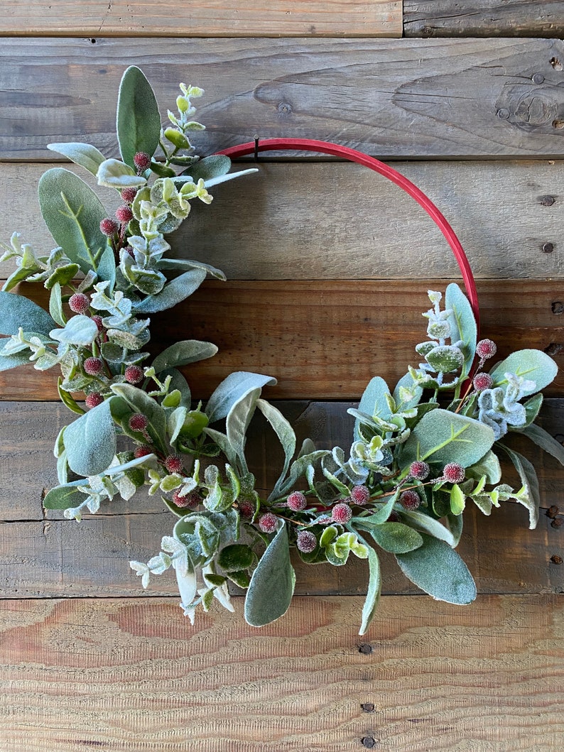 May include: A red wooden wreath with a frosted eucalyptus and berry garland. The wreath is hanging on a rustic wooden background.
