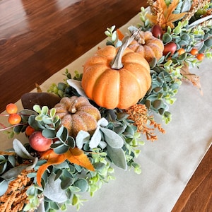 May include: A decorative table runner centrepiece featuring artificial orange pumpkins, green eucalyptus leaves, orange maple leaves, small orange berries, and textured brown accents. The arrangement is composed of various faux botanical elements.