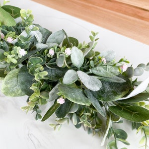 Puede incluir: Una guirnalda de eucalipto verde con peque&ntilde;as flores rosas. La guirnalda est&aacute; sobre una superficie de m&aacute;rmol blanco.