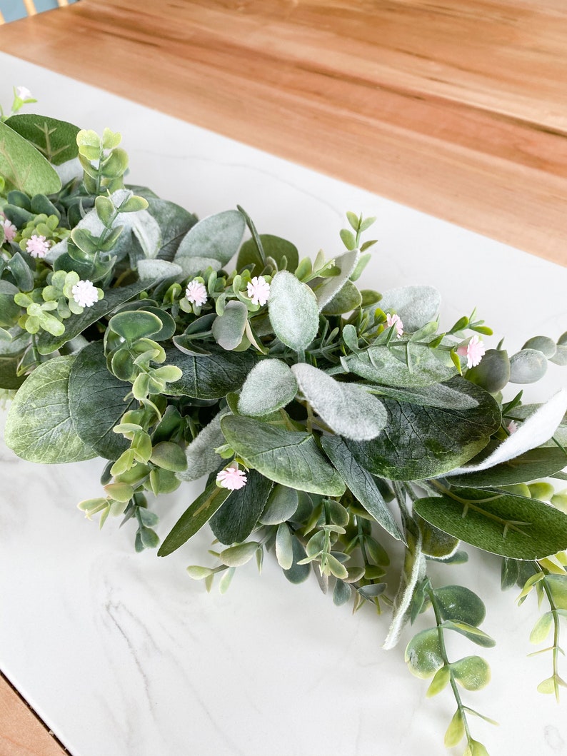 Puede incluir: Una guirnalda de eucalipto verde con peque&ntilde;as flores rosas. La guirnalda est&aacute; sobre una superficie de m&aacute;rmol blanco.