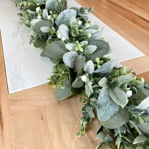 Puede incluir: Una guirnalda verde de eucalipto y vegetaci&oacute;n con flores blancas, perfecta para una decoraci&oacute;n de mesa.