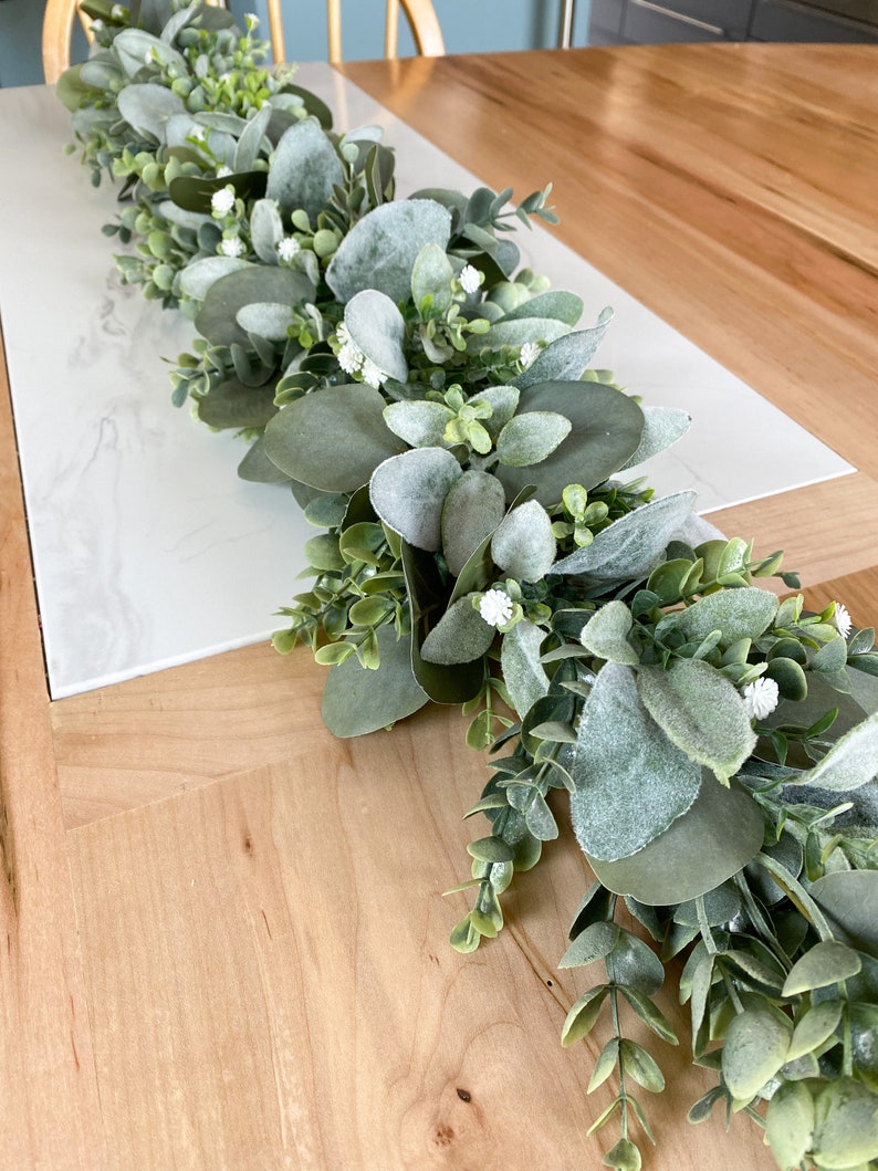 Puede incluir: Una guirnalda verde de eucalipto y vegetaci&oacute;n con flores blancas, perfecta para una decoraci&oacute;n de mesa.