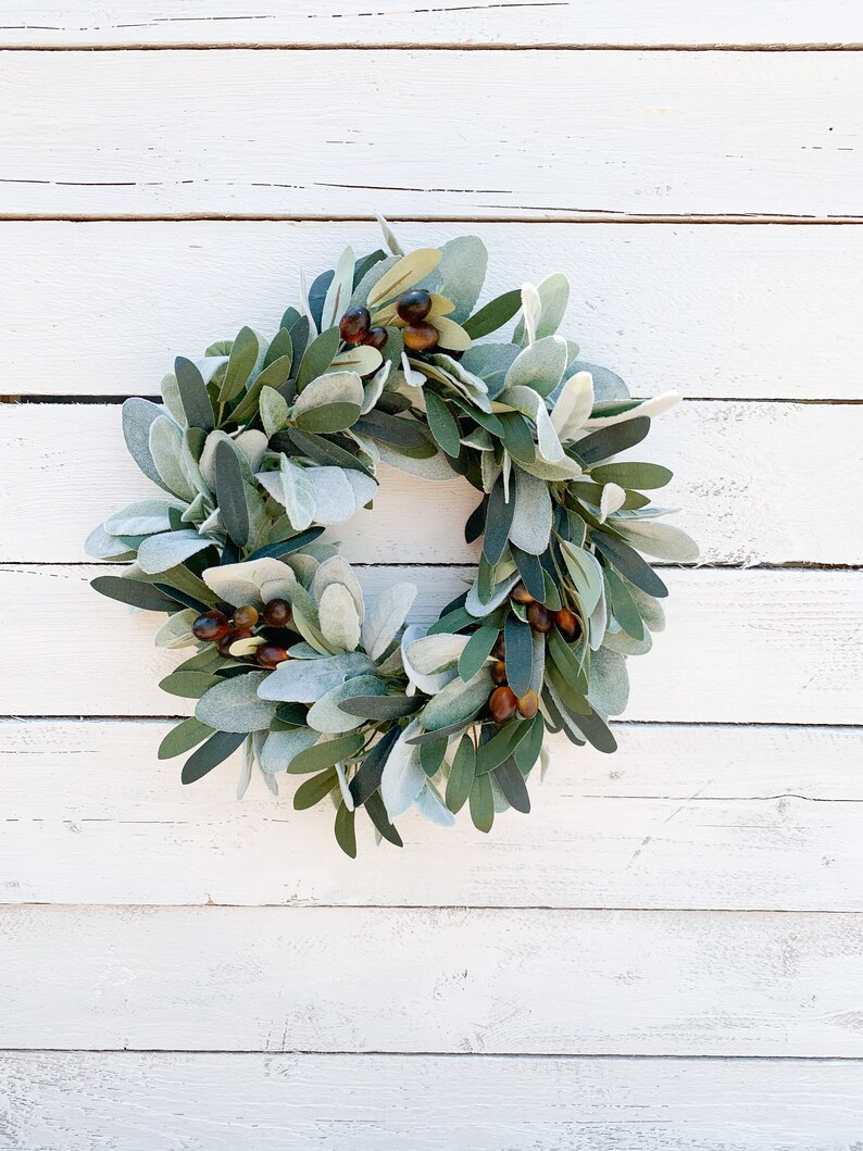 May include: A green and brown wreath made of faux greenery and berries. The wreath is hanging on a white wooden wall.