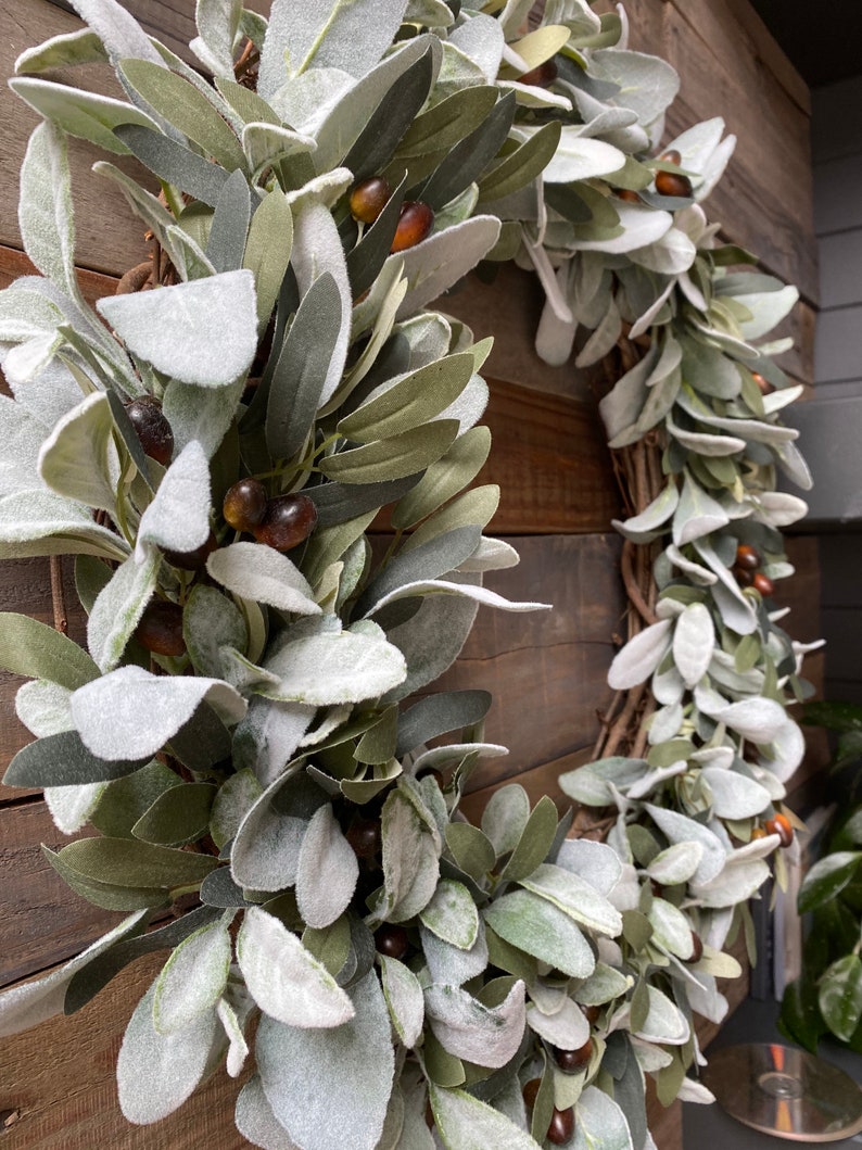 May include: A green and white wreath made of faux lamb's ear leaves with brown berries. The wreath is hanging on a wooden wall.