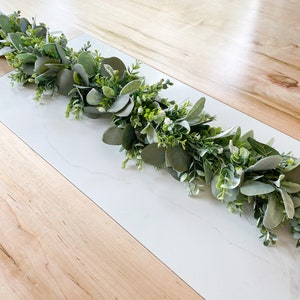 Puede incluir: Una guirnalda verde de eucalipto y vegetaci&oacute;n con peque&ntilde;as flores blancas. La guirnalda est&aacute; sobre una superficie de madera clara con una superficie de m&aacute;rmol blanco.