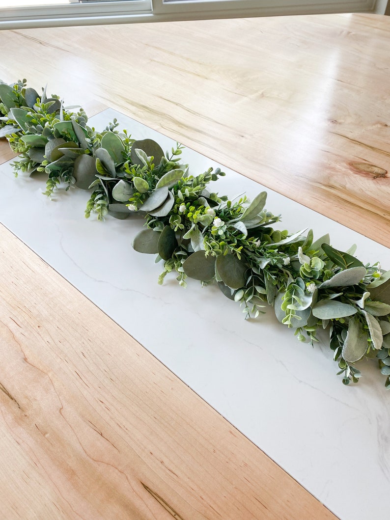 Puede incluir: Una guirnalda verde de eucalipto y vegetaci&oacute;n con peque&ntilde;as flores blancas. La guirnalda est&aacute; sobre una superficie de madera clara con una superficie de m&aacute;rmol blanco.