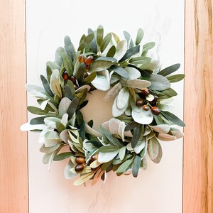 May include: A green and brown olive branch wreath with faux berries. The wreath is made of artificial greenery and is perfect for adding a touch of nature to your home decor.