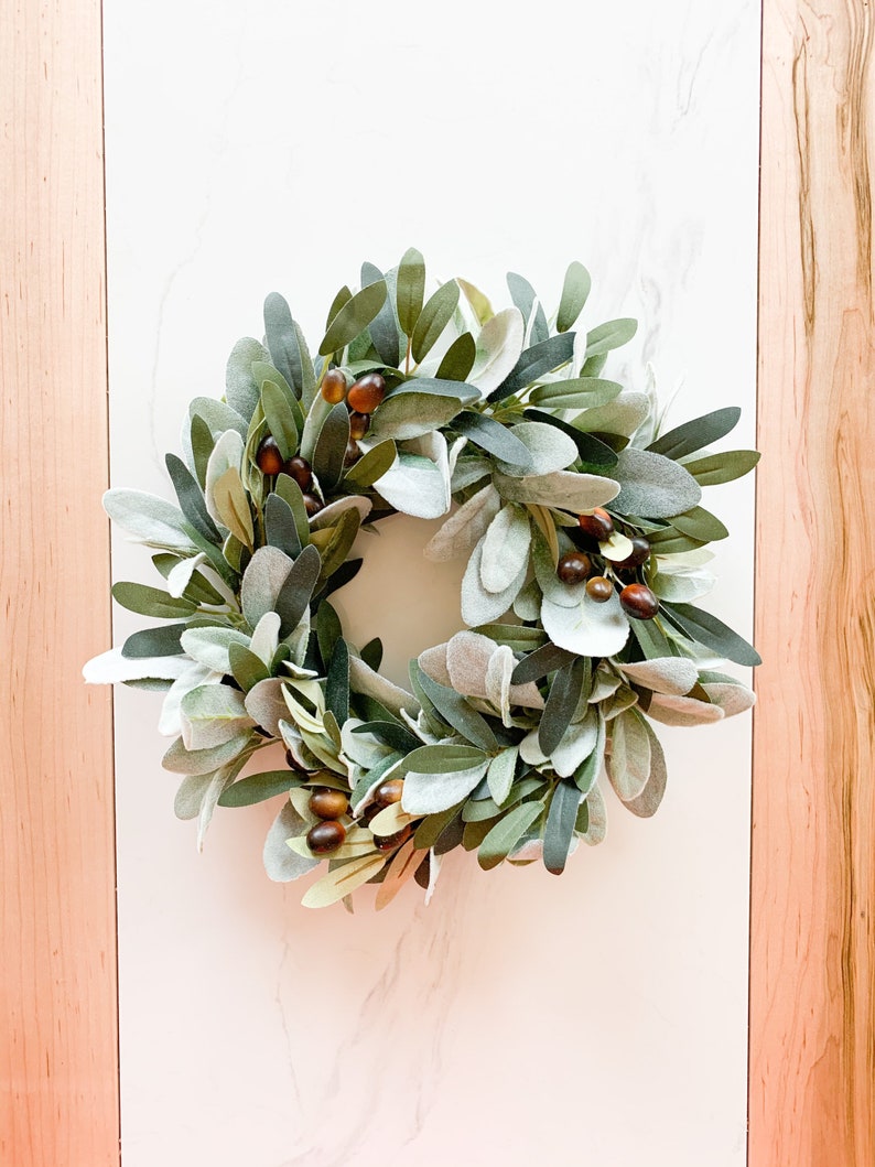 May include: A green and brown olive branch wreath with faux berries. The wreath is made of artificial greenery and is perfect for adding a touch of nature to your home decor.