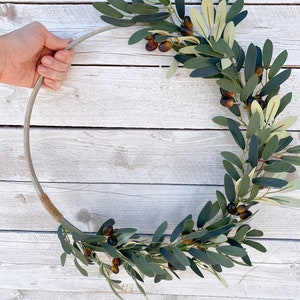 May include: A green olive branch wreath with brown olives, wrapped around a silver metal ring. The wreath is on a white wooden background.