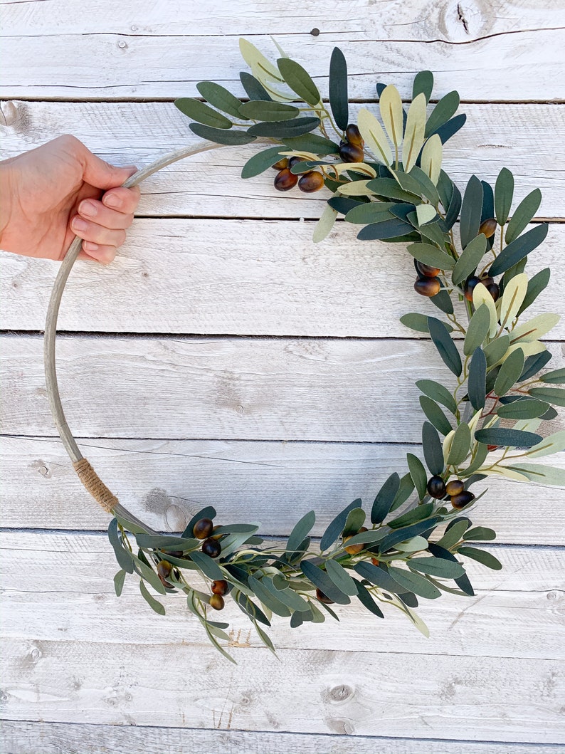 May include: A green olive branch wreath with brown olives, wrapped around a silver metal ring. The wreath is on a white wooden background.