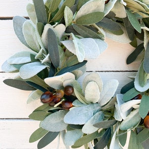 May include: A green and gray artificial wreath with brown berries. The wreath is made of faux leaves and berries.