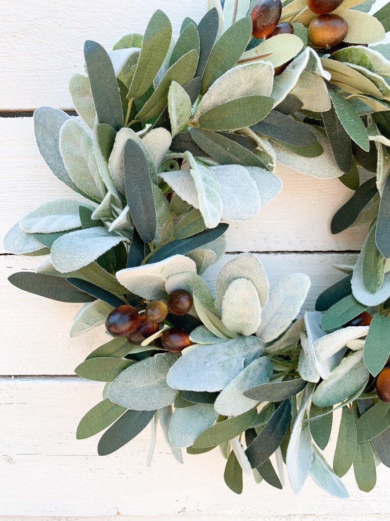 May include: A green and gray artificial wreath with brown berries. The wreath is made of faux leaves and berries.