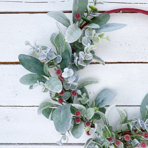 May include: A frosted eucalyptus garland with red berries. The garland is on a white wooden background.