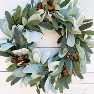 May include: A green and brown olive branch wreath with faux olives. The wreath is made of artificial greenery and is perfect for adding a touch of nature to your home decor.