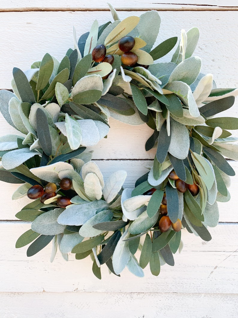 May include: A green and brown olive branch wreath with faux olives. The wreath is made of artificial greenery and is perfect for adding a touch of nature to your home decor.