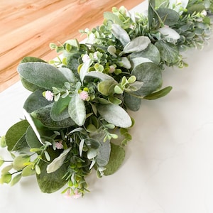 Puede incluir: Una guirnalda de eucalipto verde y blanca con peque&ntilde;as flores rosas. La guirnalda est&aacute; sobre una superficie blanca.