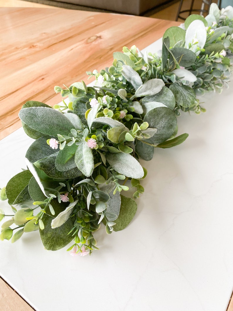Puede incluir: Una guirnalda de eucalipto verde y blanca con peque&ntilde;as flores rosas. La guirnalda est&aacute; sobre una superficie blanca.