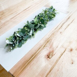 Puede incluir: Un camino de mesa blanco y de madera con una guirnalda verde de hojas y peque&ntilde;as flores blancas.