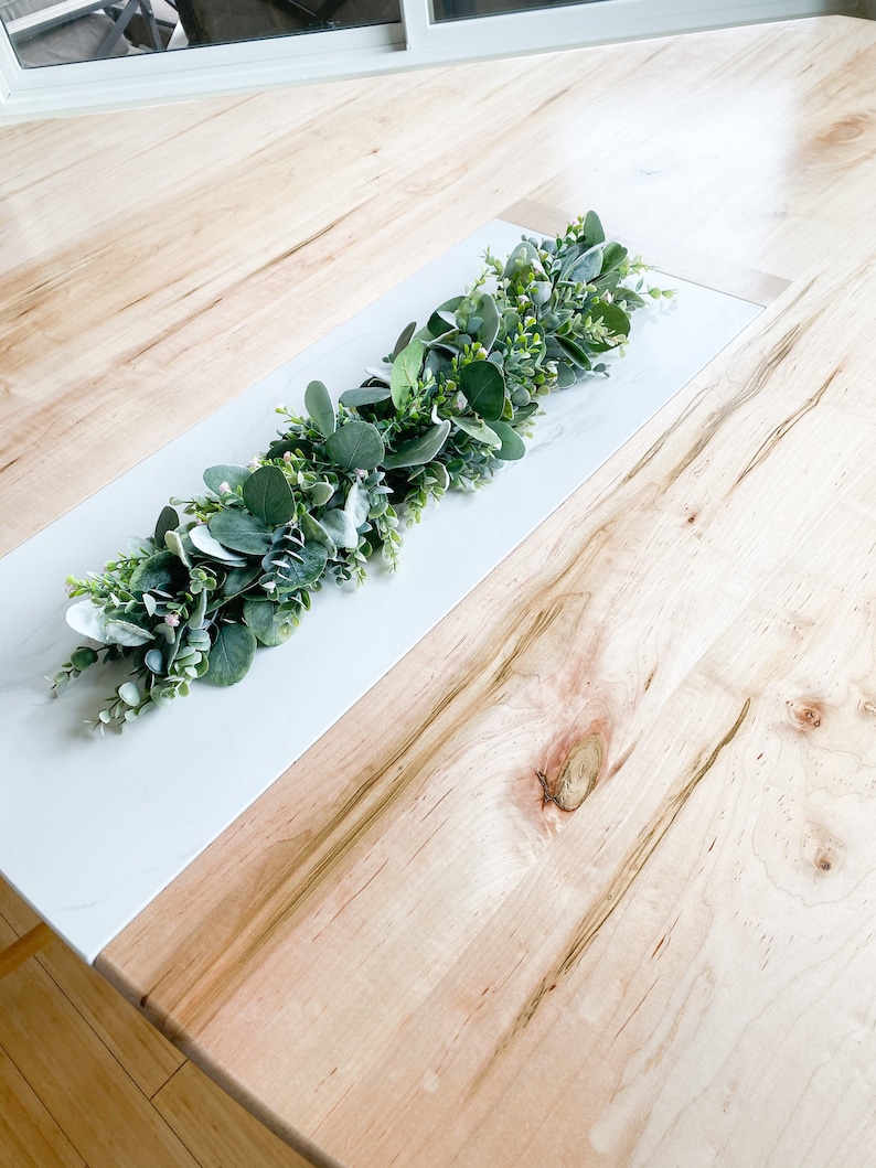 Puede incluir: Un camino de mesa blanco y de madera con una guirnalda verde de hojas y peque&ntilde;as flores blancas.