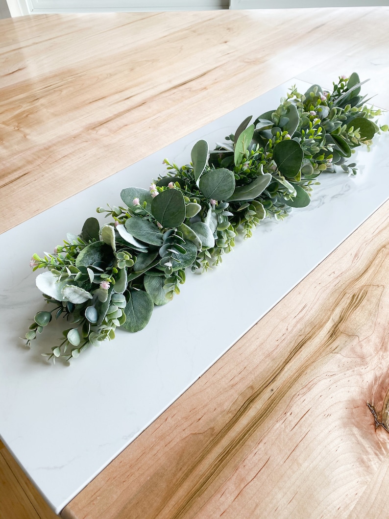 Puede incluir: Una guirnalda de eucalipto verde y blanca con peque&ntilde;as flores rosas, ideal para decorar una mesa o una repisa de la chimenea.