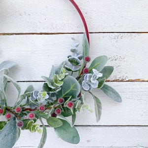 May include: A red wooden hoop wreath with frosted eucalyptus leaves and red berries. The wreath is on a white wooden background.