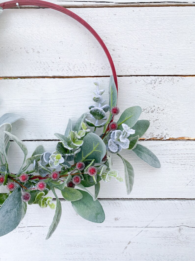May include: A red wooden hoop wreath with frosted eucalyptus leaves and red berries. The wreath is on a white wooden background.