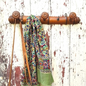 May include: A wooden coat rack with three hooks, mounted on a white painted wall. A green and blue patterned scarf and a brown leather purse hang from the hooks.