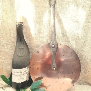 May include: A vintage copper pan with a long, metal handle. The pan has a round, flat bottom and is slightly dented. The pan is sitting on a burlap surface next to a bottle of wine.
