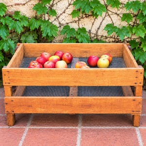May include: A rectangular wooden crate with a wire mesh bottom, containing a collection of red and yellow apples. The crate is constructed from light brown wood planks and features a sturdy frame with four short legs, designed for storage.