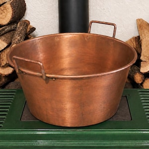 May include: A large, round, hammered copper basin with a wide, open top and two rectangular metal handles attached to the sides. The basin features a smooth, metallic finish with visible signs of wear and a warm, reddish-brown colour throughout.