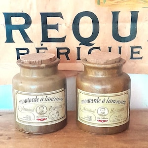 Puede incluir: Dos tarros de mostaza antiguos con tapones de corcho. Cada tarro tiene una etiqueta con el texto "moutarde à l'ancienne Armand Bizonand" y el logotipo de AMORA. Los tarros son de color marrón claro y están sobre una superficie de madera.