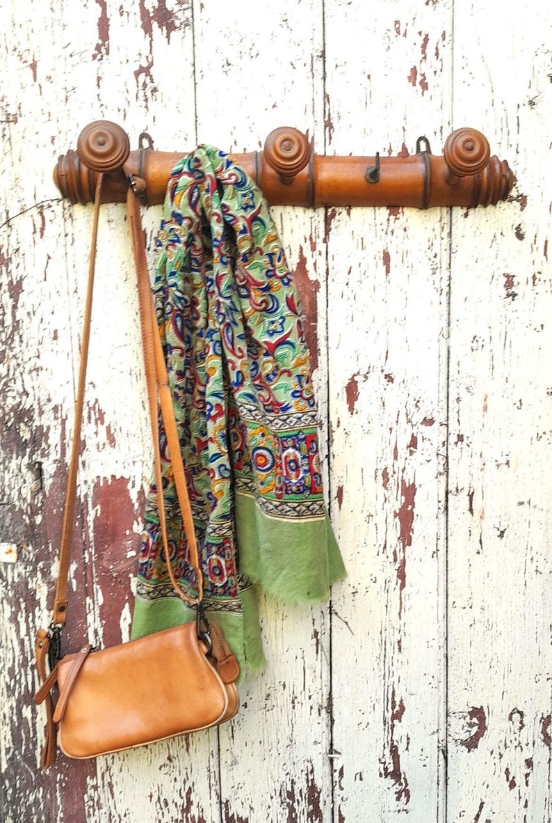 May include: A wooden coat rack with three hooks, mounted on a white painted wall. A green and blue patterned scarf and a brown leather purse hang from the hooks.