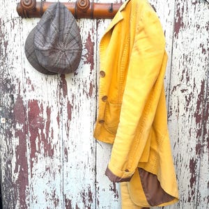 May include: A yellow corduroy jacket with brown trim hangs on a wooden coat rack with a brown and gray tweed hat.