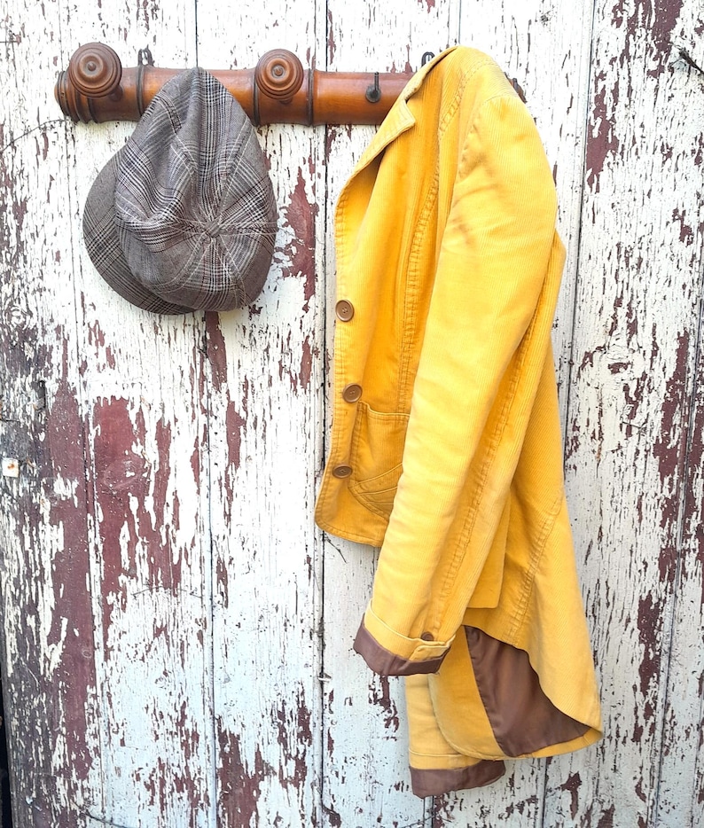May include: A yellow corduroy jacket with brown trim hangs on a wooden coat rack with a brown and gray tweed hat.