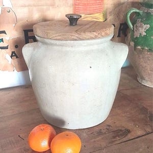 Könnte beinhalten: Ein cremefarbener Keramiktopf mit Holzdeckel und dunklem Knopf. Zwei Orangen liegen vor dem Topf auf einer Holzoberfläche. Der Topf hat zwei kleine Griffe an den Seiten.
