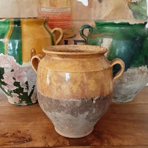 May include: A selection of antique ceramic pots in shades of yellow, brown, and green. The pots have a weathered appearance with visible imperfections, suggesting age and use. Each pot features a rounded body and handles, with a rustic aesthetic.