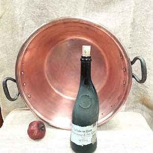 Puede incluir: Una olla grande de cobre con dos asas de hierro negro. La olla está sentada sobre una superficie de arpillera con una botella de vino y una manzana roja delante de ella. La etiqueta de la botella de vino dice "La Jolie du Pape Chateauneuf-du-Pape."