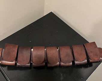 May include: A brown leather belt with eight pouches, each with a rounded flap. The belt is lying on a black surface.