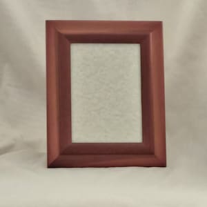 May include: A rectangular, reddish-brown wooden picture frame. The frame has a simple, classic design with a slightly beveled edge. The interior space is empty, with a light-colored, textured backing.