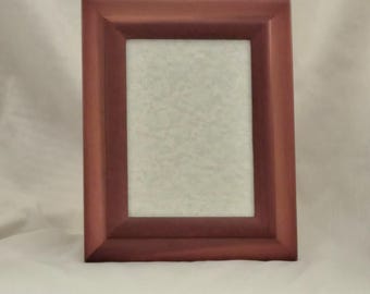 May include: A rectangular, reddish-brown wooden picture frame. The frame has a simple, classic design with a slightly beveled edge. The interior space is empty, with a light-colored, textured backing.
